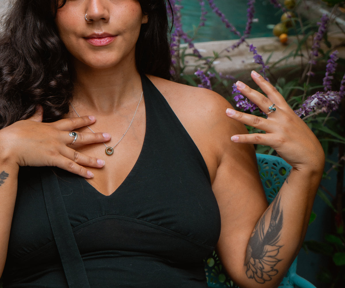 A woman wearing a delicate silver chain necklace with a small circular pendant, styled with silver rings on her fingers, in an outdoor garden setting with purple flowers.