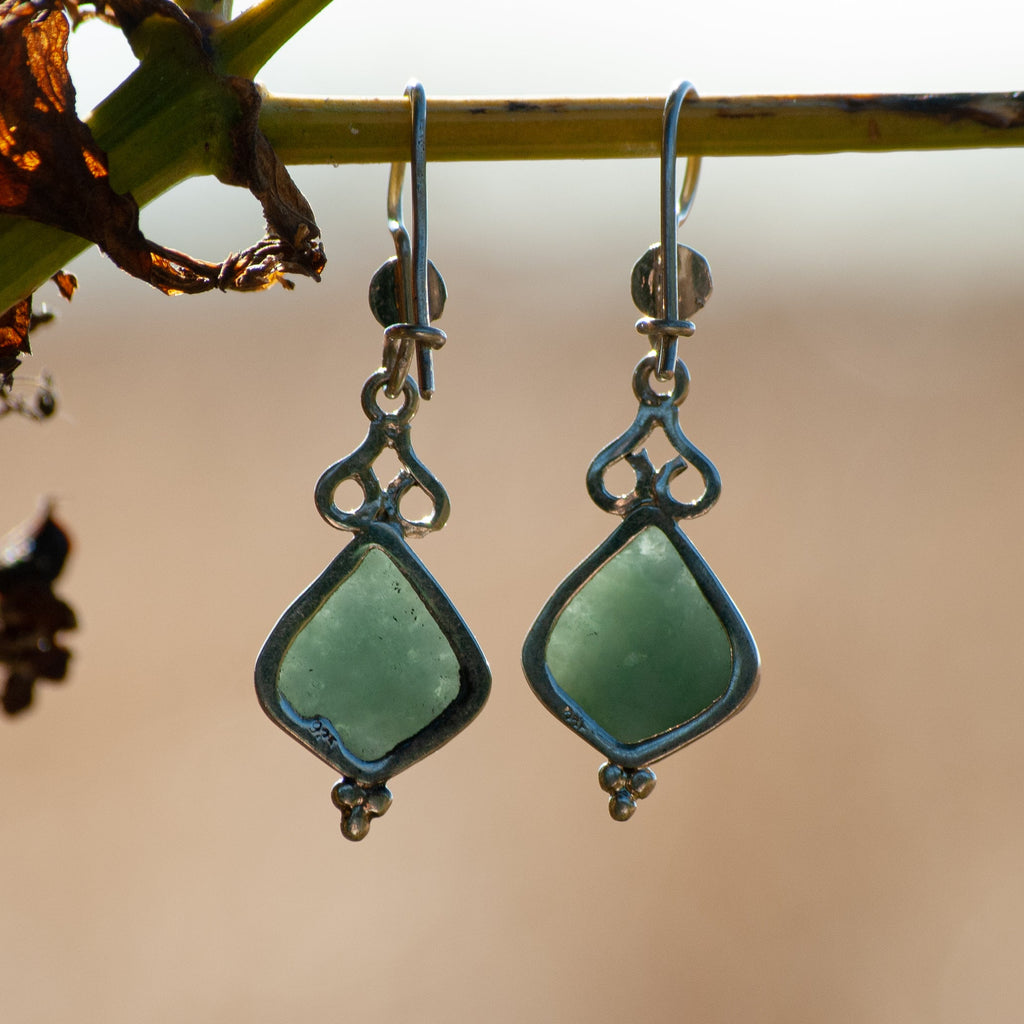 A pair of dangling earrings with light green teardrop-shaped stones in silver-toned settings, hanging from a thin branch against a soft beige background.