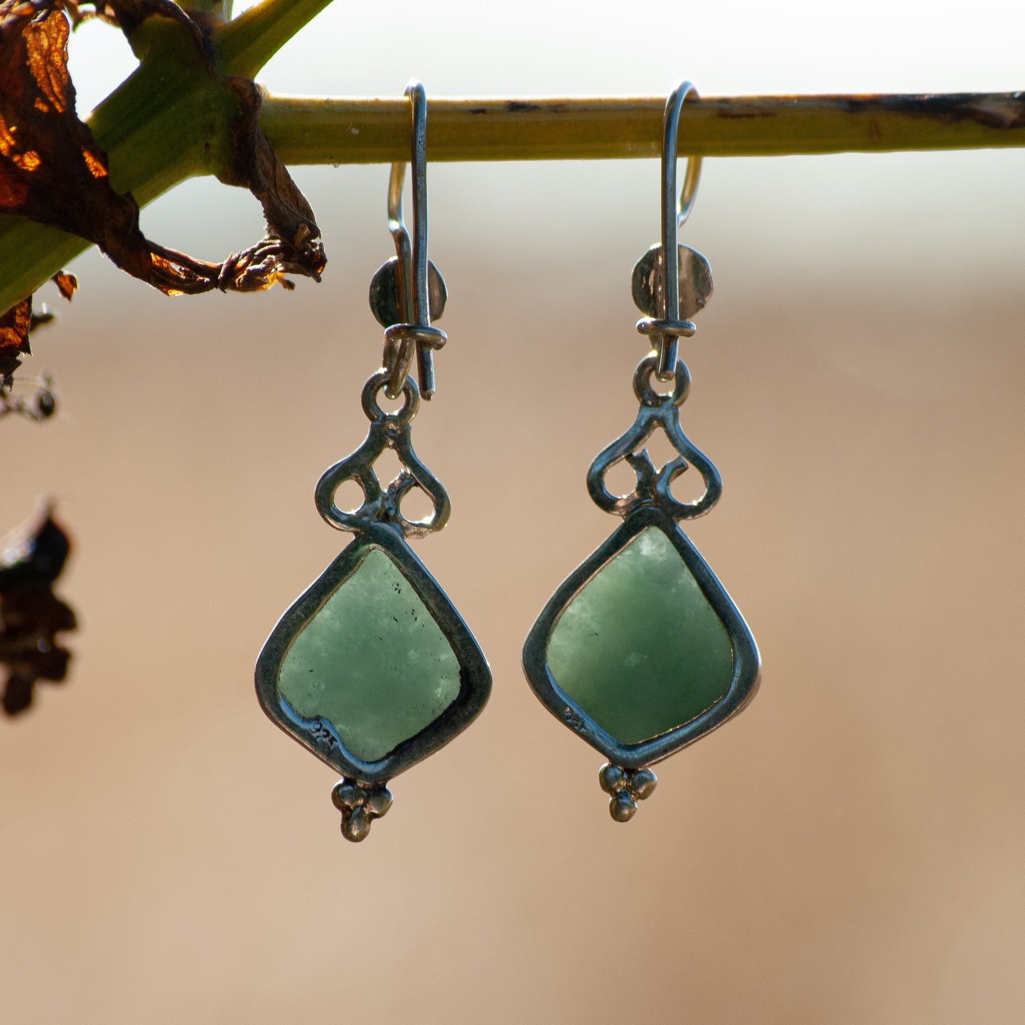 A pair of dangling earrings with light green teardrop-shaped stones in silver-toned settings, hanging from a thin branch against a soft beige background.