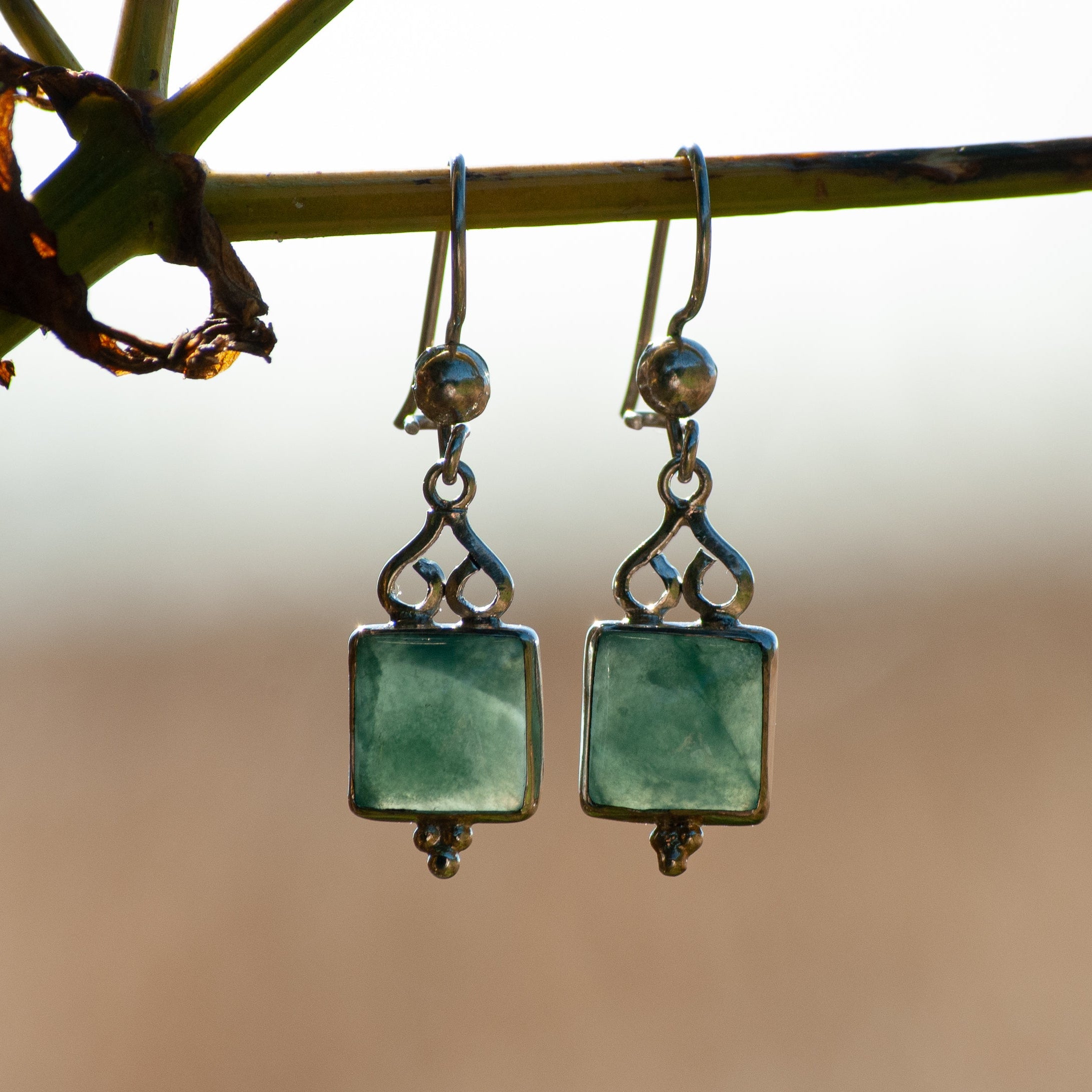 A pair of silver drop earrings with square green gemstone pendants, hanging from a plant stem against a soft, blurred beige background.