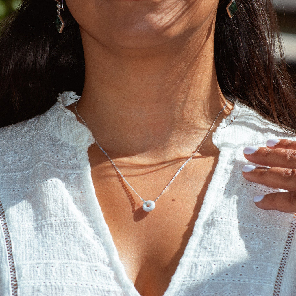 A close-up lifestyle shot of a woman wearing a delicate silver double-layer necklace with a small white jade pendant, styled with silver drop earrings and a silver ring on her finger, set against a softly blurred outdoor background.