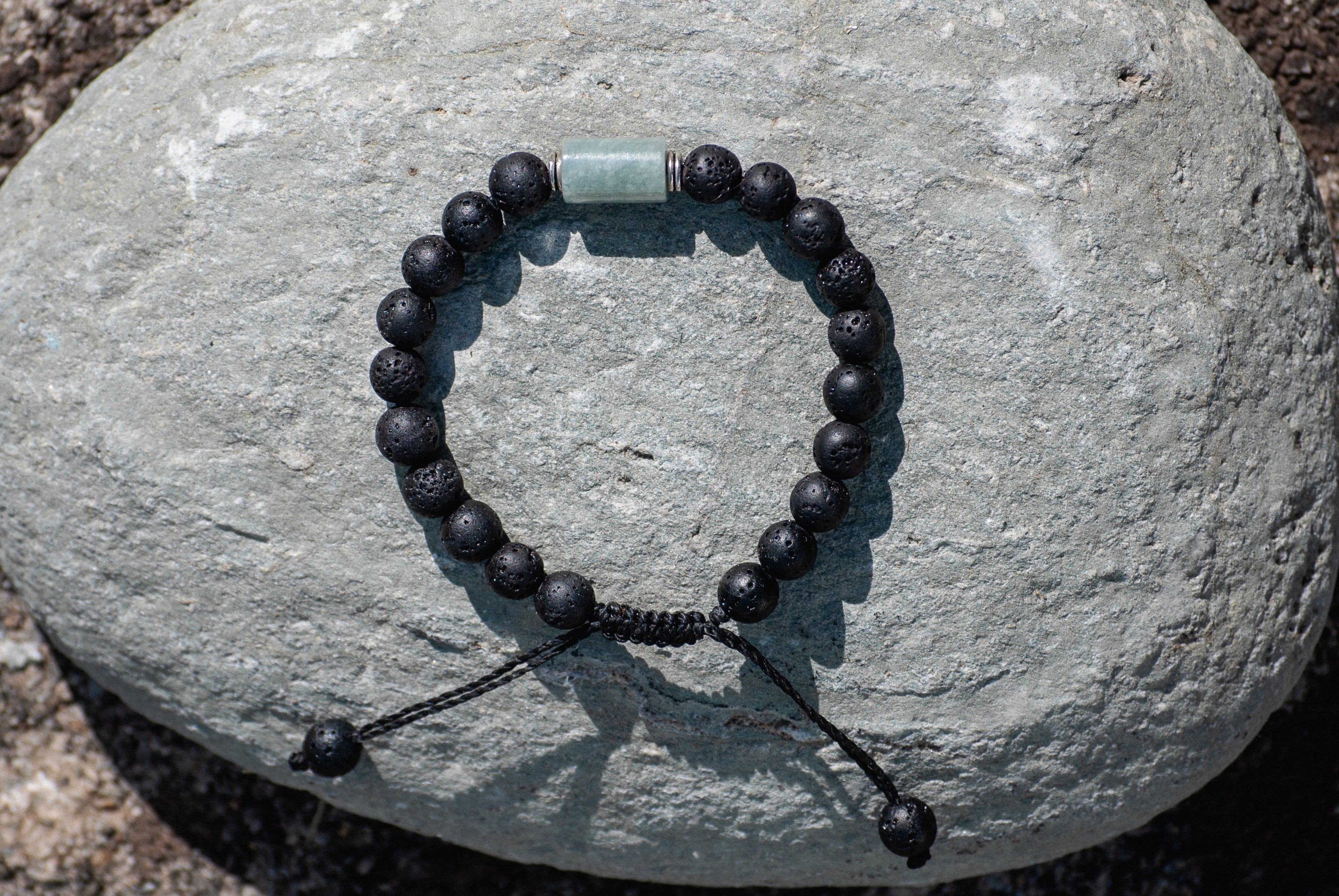 A beaded bracelet with matte black lava stone beads and light green jade cylinder, displayed on a smooth grey rock outdoors.
