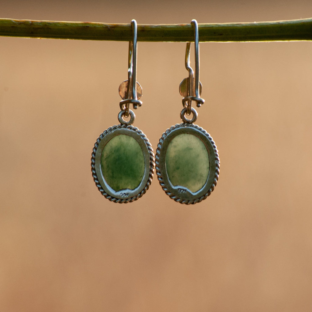 A pair of silver-toned dangle earrings with oval green cabochon stones, hanging from a thin green twig against a warm, blurred tan background.