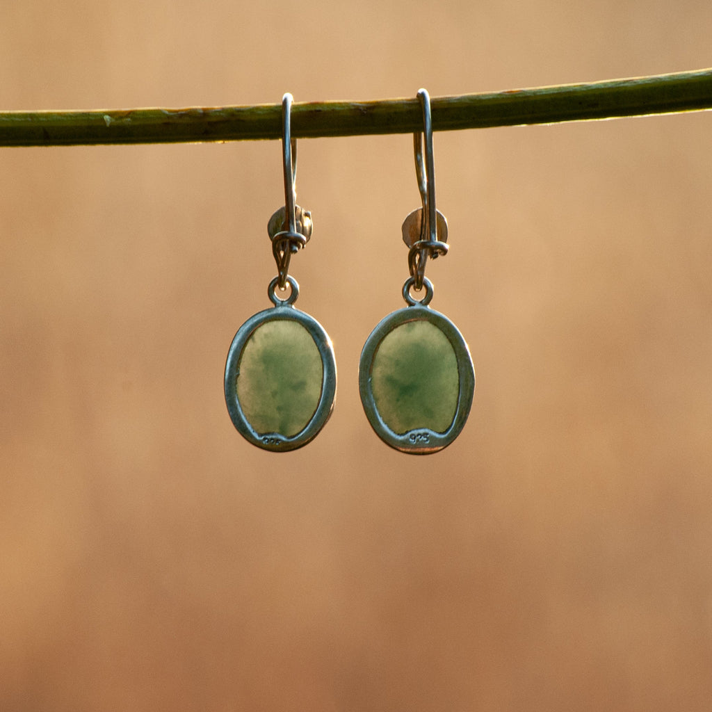 A pair of silver drop earrings with oval light green stone pendants, hanging from a green twig against a warm, blurred brown background.