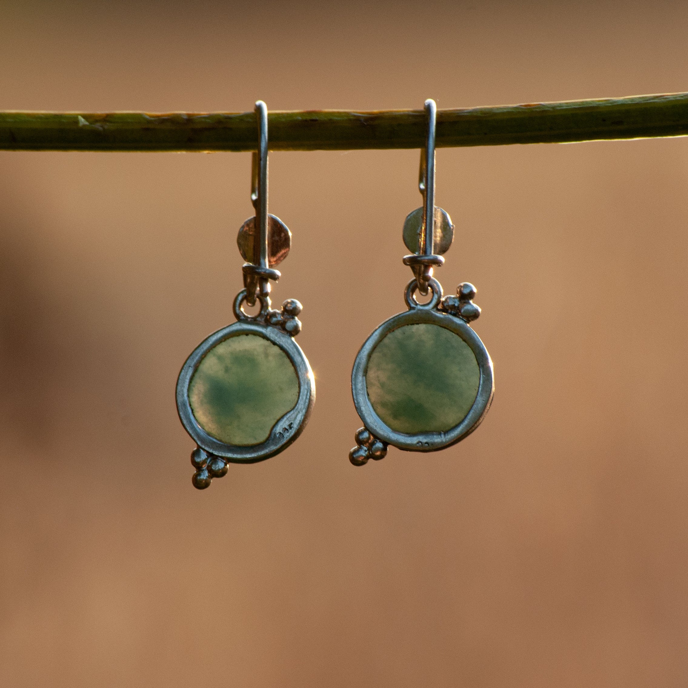 A pair of silver-toned dangle earrings with oval light green stone inlays, hanging from a green twig against a warm, blurred brown background.