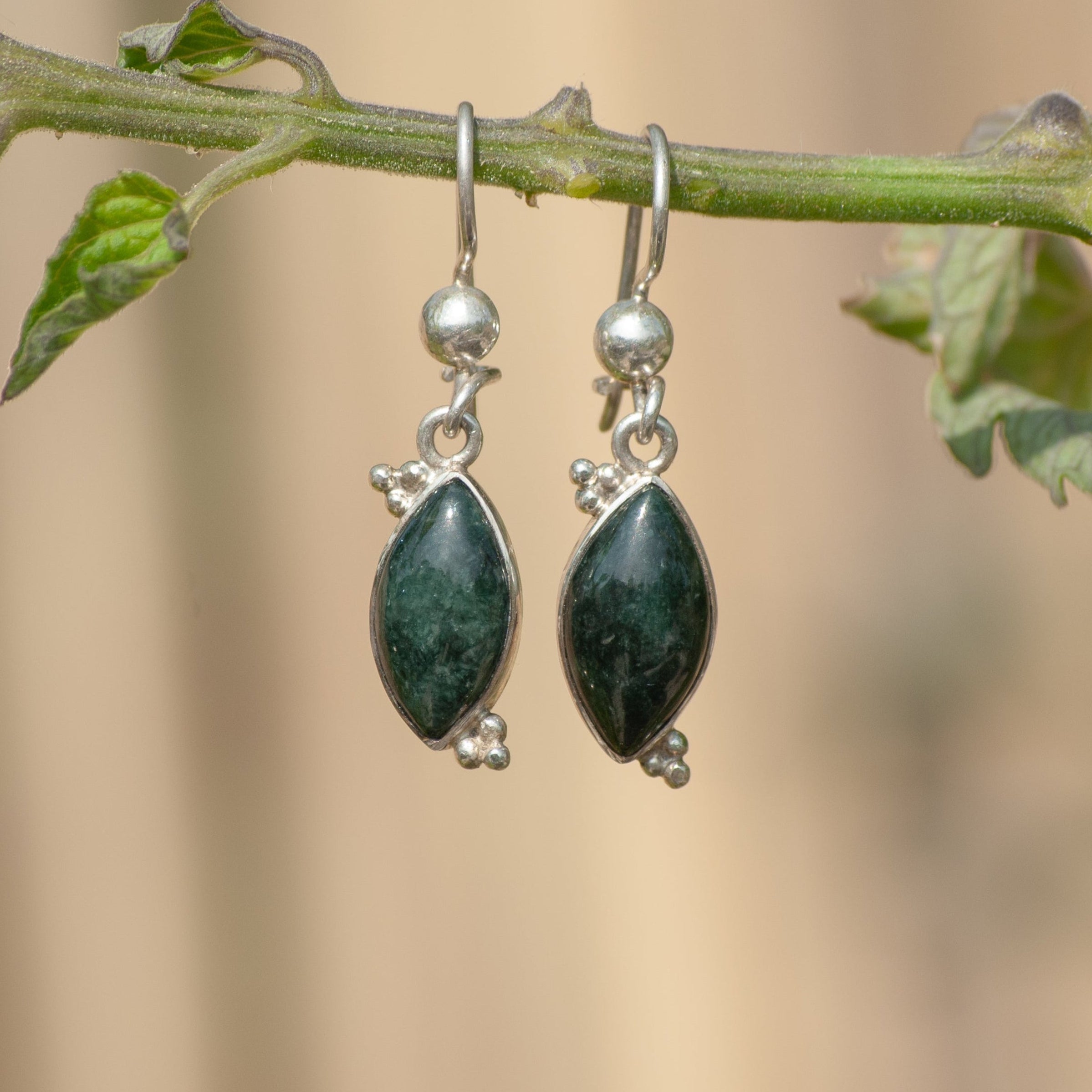A pair of silver dangle earrings with marquise-shaped dark green gemstones hanging from a green plant stem against a soft beige background.
