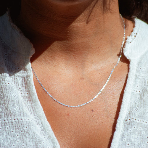 A close-up of a person wearing a delicate silver chain necklace over a white eyelet blouse, with bright outdoor sunlight casting shadows.