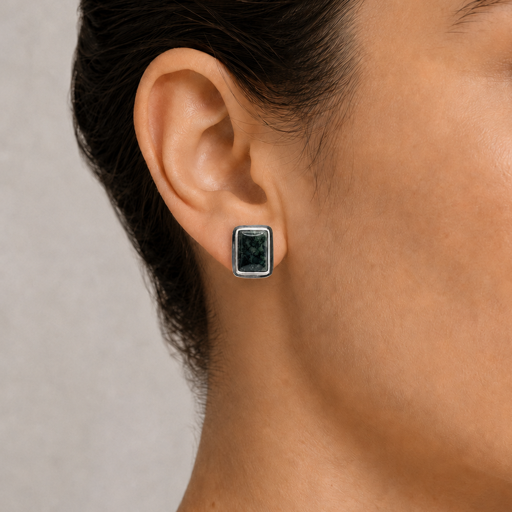 Close-up of an ear wearing a square-shaped earring with a dark jade gemstone on a neutral background