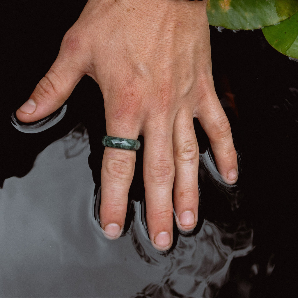 A close-up lifestyle shot of a person's hand resting in dark water next to green lily pads. The person is wearing a dark beaded bracelet with a light blue stone and a dark green ring.