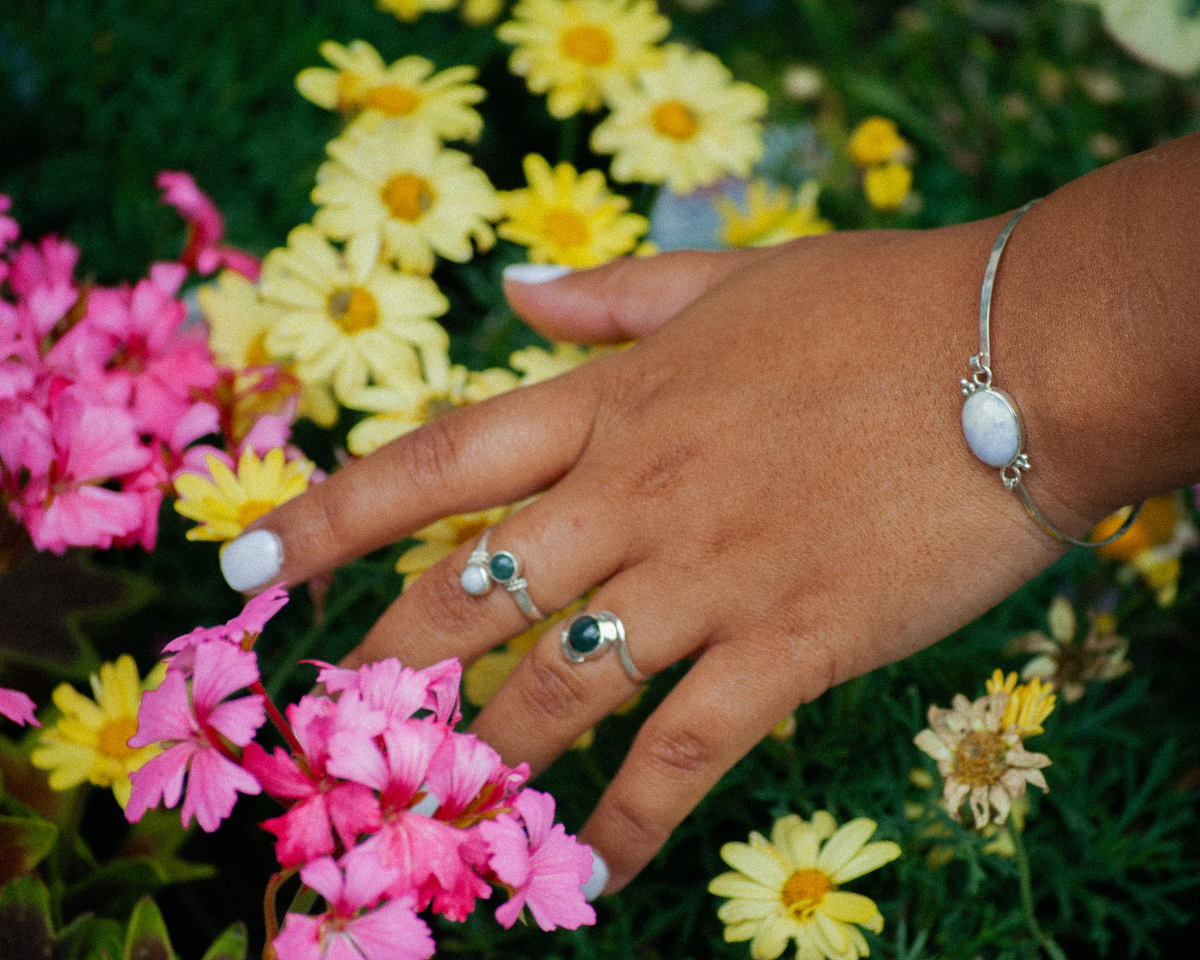 A close-up of a hand wearing silver rings and a silver bracelet with white stones, set against a background of pink and yellow flowers.