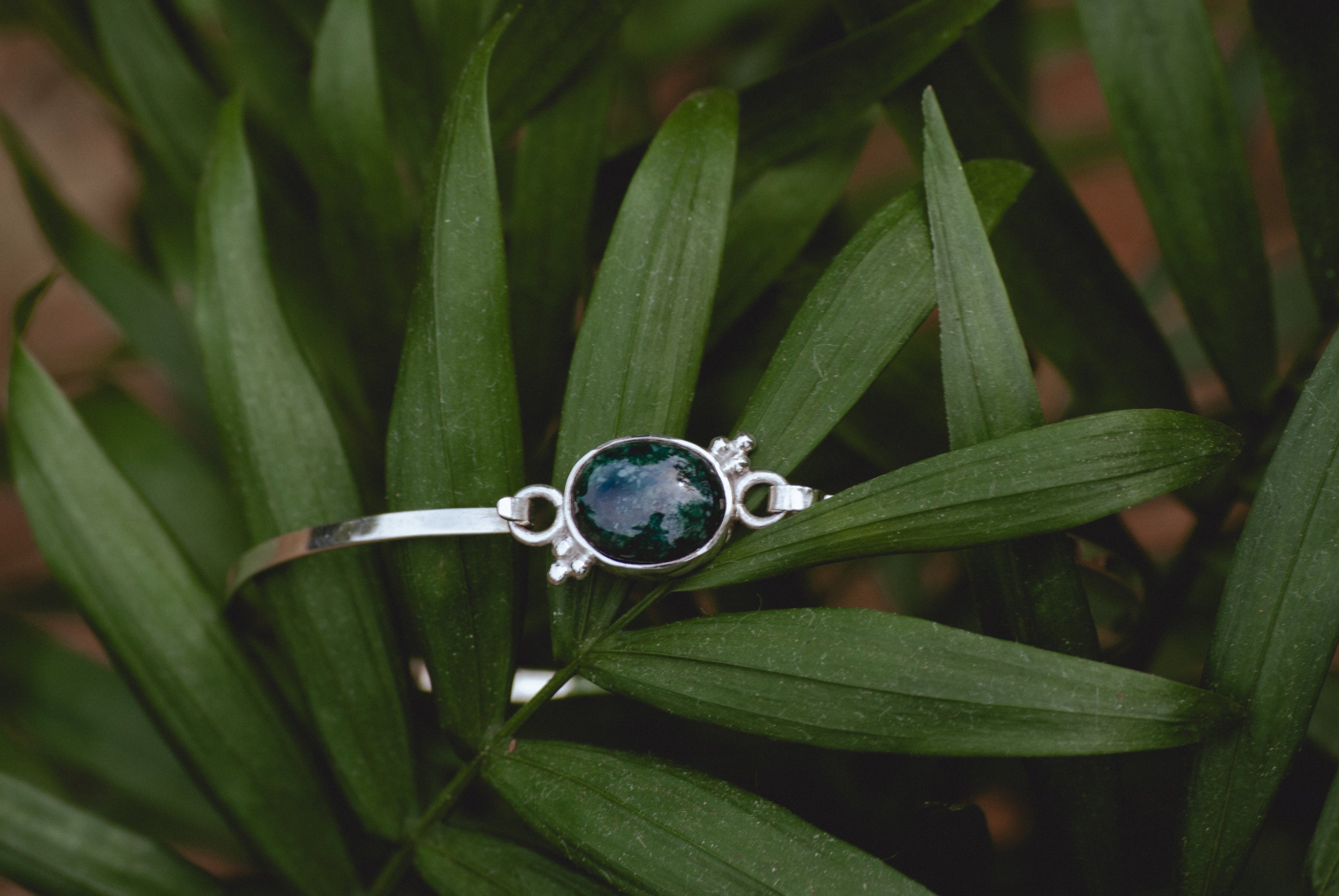 A silver bangle bracelet with a central oval dark green stone and decorative side details, resting on green palm leaves.