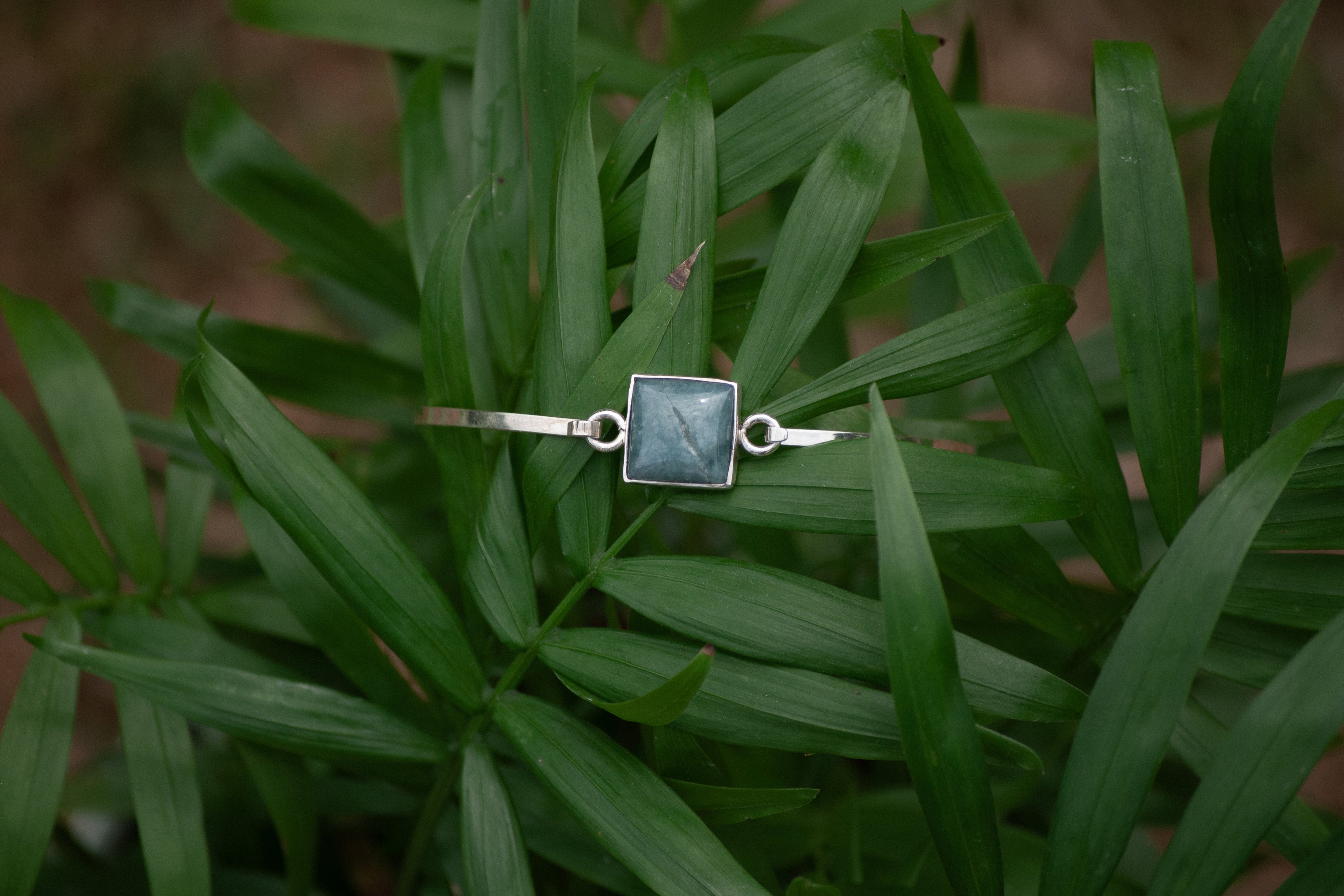 A silver-toned ring with a square dark green stone, resting on a bed of green palm leaves.