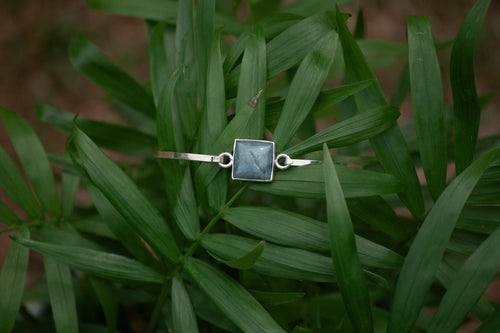 A silver-toned ring with a square dark green stone, resting on a bed of green palm leaves.