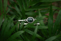A silver-toned bangle bracelet with a round dark green jade stone, resting on a bed of green palm leaves.