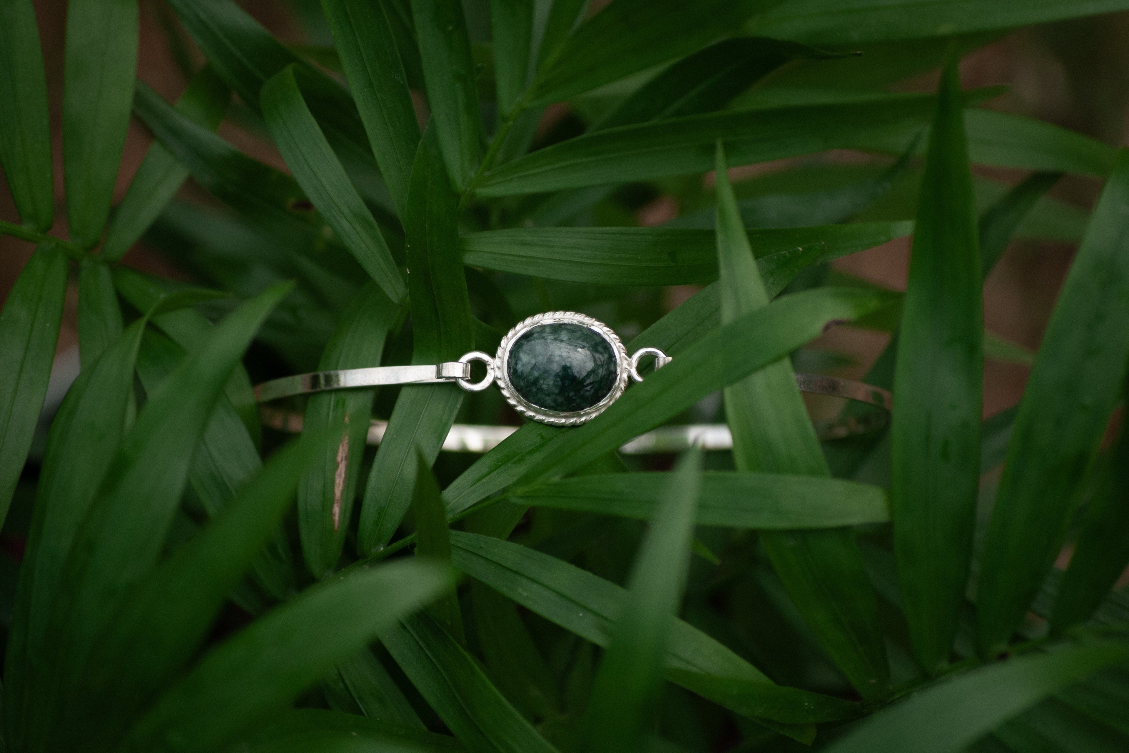 A silver-toned bangle bracelet with a round dark green jade stone, resting on a bed of green palm leaves.