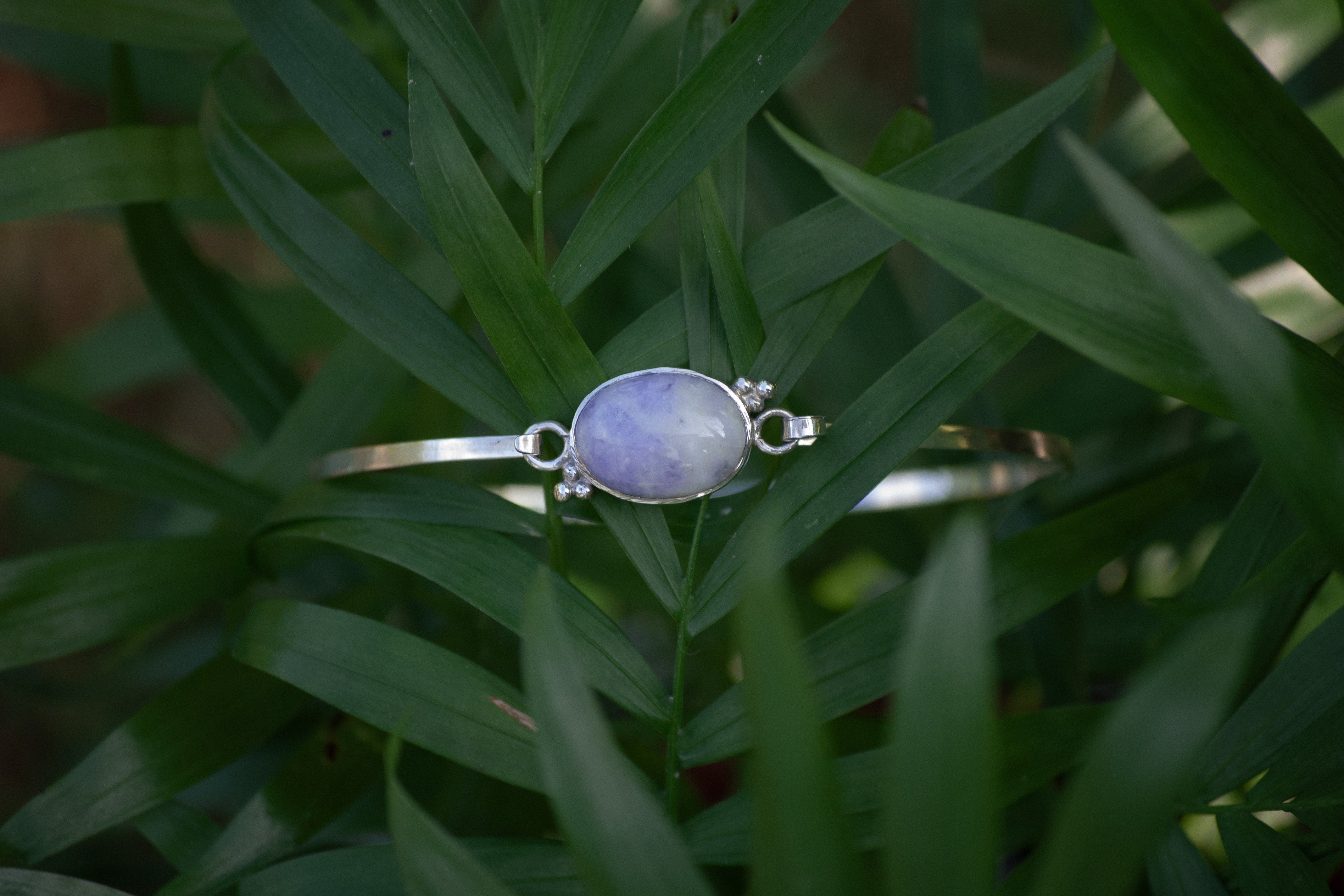 A silver bangle bracelet with an oval light purple stone, resting on green leaves.