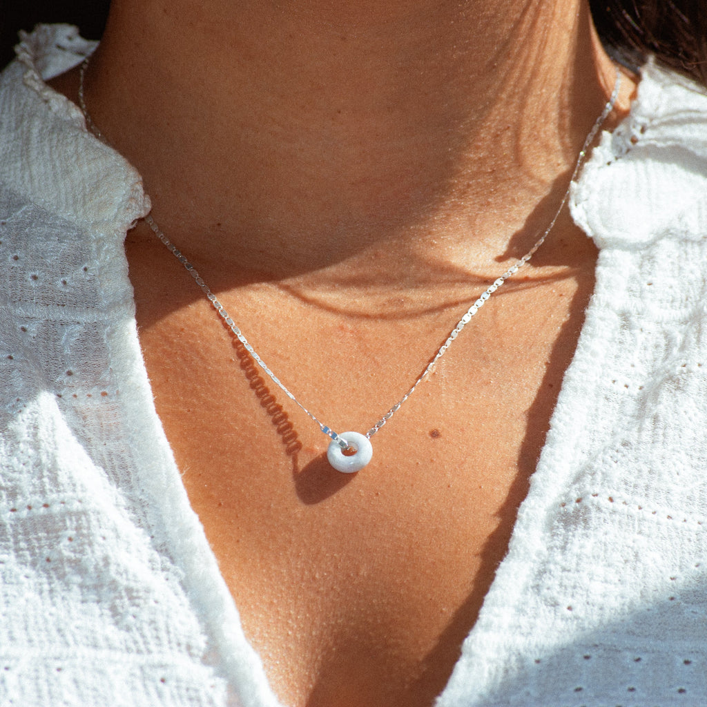 A close-up of a person wearing a delicate silver chain necklace with a small white circular jade pendant, styled with a white textured eyelet top.