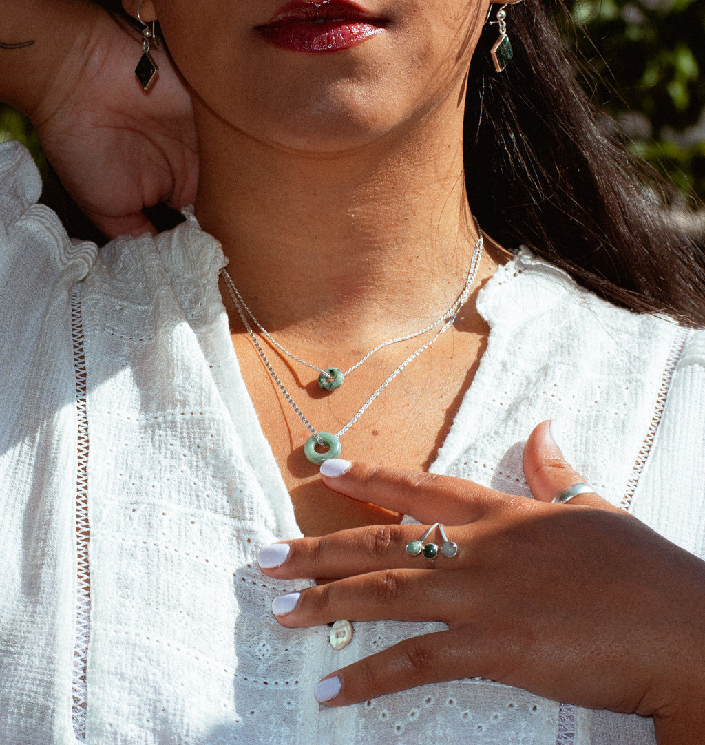 A close-up lifestyle shot of a woman wearing layered silver necklaces with green stone pendants and matching green earrings, styled with a white eyelet blouse.