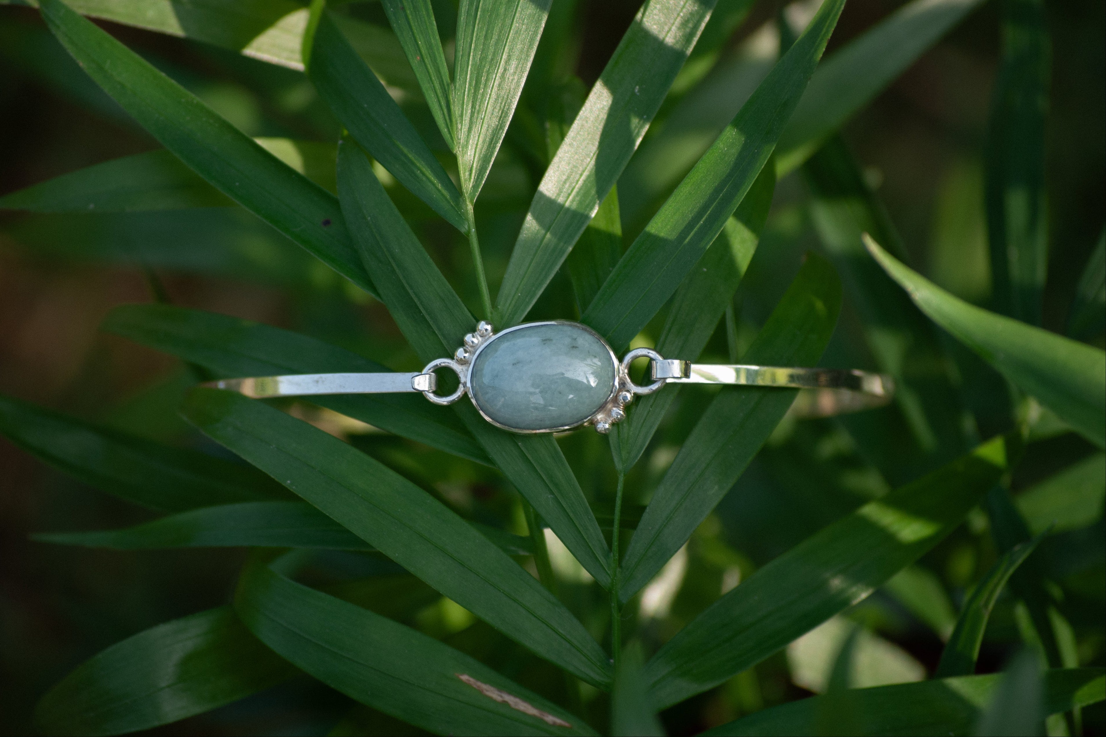 A silver-toned bangle bracelet with a central oval light blue-green stone and small clear stones on either side, resting on green palm leaves.