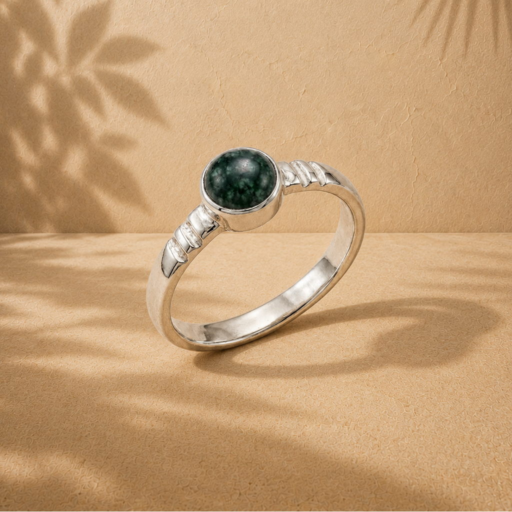 A silver ring with a round dark green jade stone, displayed on a textured beige surface with leaf shadows cast across it.