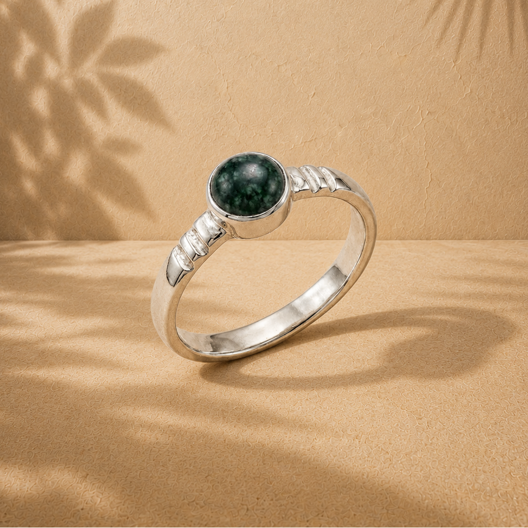 A silver ring with a round dark green jade stone, displayed on a textured beige surface with leaf shadows cast across it.