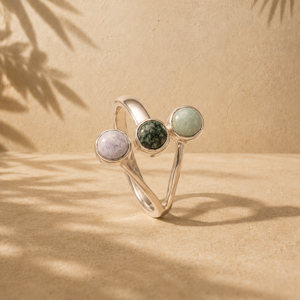 A silver-toned open ring with three round bezel-set jade gemstones in pale lavender, dark green, and light green, displayed on a sandy beige surface with palm leaf shadows.