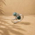 A silver ring with two round green jade gemstone settings, displayed on a textured beige surface with palm leaf shadows cast across it.