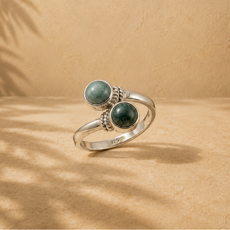 A silver ring with two round green jade gemstone settings, displayed on a textured beige surface with palm leaf shadows cast across it.